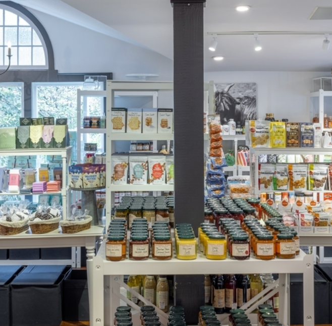 A well-lit store interior features shelves stocked with jars, bottles, packaged foods, and greeting cards. Sunlight streams through a large arched window, creating a welcoming atmosphere.