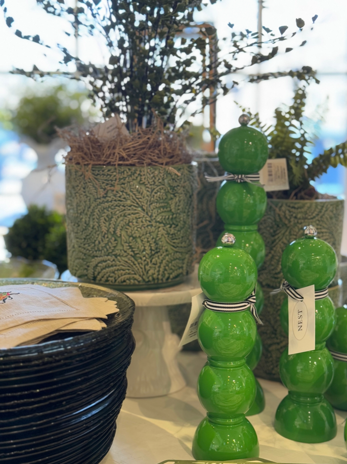 A display featuring stacked green decorative objects, leafy potted plants in textured pots, and a stack of black plates on a table. Some items are labeled with tags and ribbon.