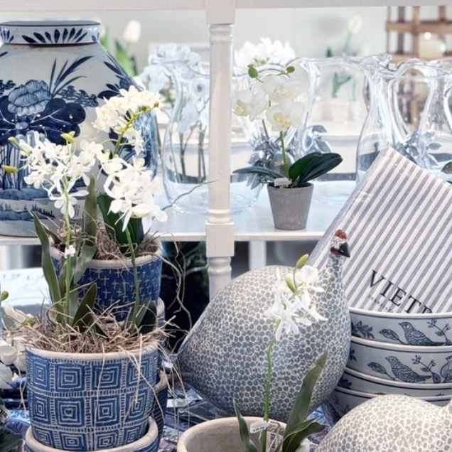 A display of blue and white ceramic vases, patterned bowls, glass pitchers, artificial white orchids, and two decorative bird figurines arranged on a table.