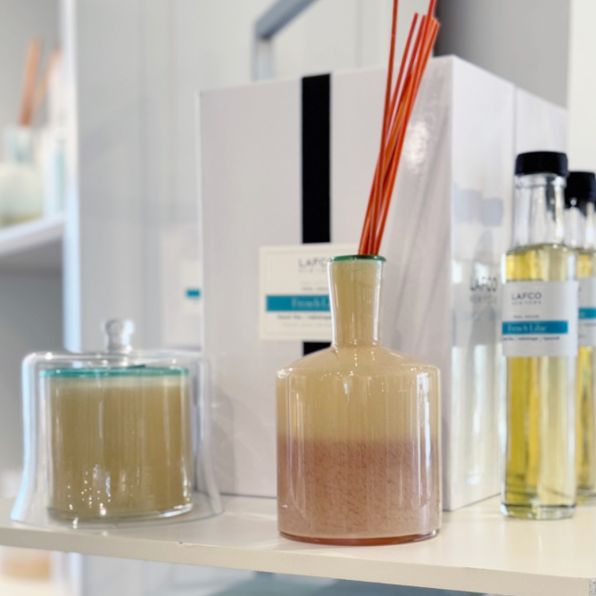 A glass diffuser with orange reeds, a lidded candle, and bottles of fragrance oil are displayed on a white shelf alongside a white box with a blue label. The setting appears to be a home or store.