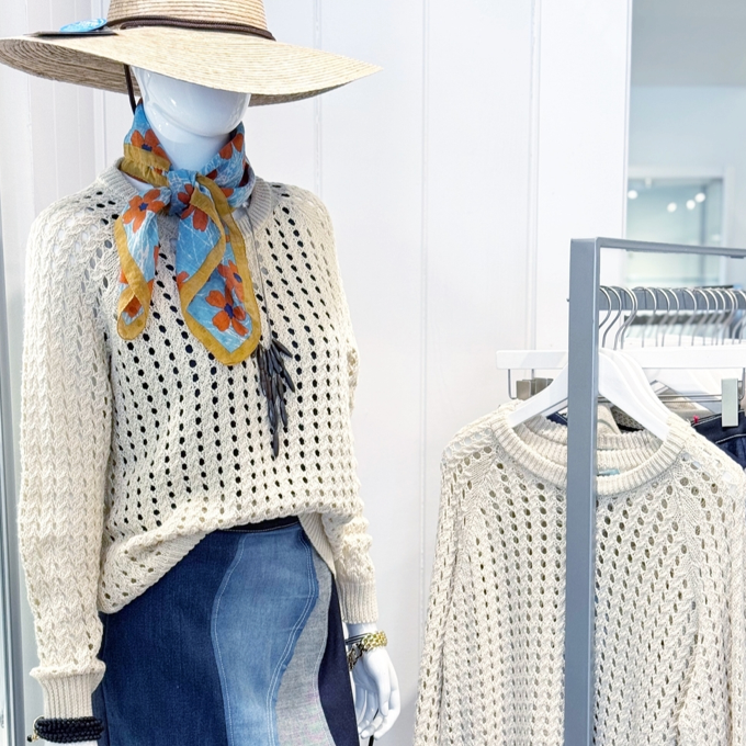 A mannequin dressed in a cream knit sweater, blue patchwork skirt, colorful floral scarf, and wide-brim straw hat stands next to a matching sweater on a hanger in a bright, modern clothing store.