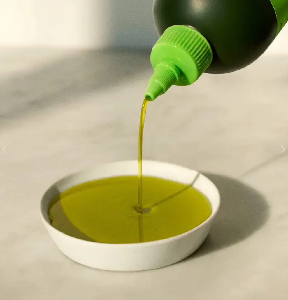 A green-tipped squeeze bottle pours golden olive oil into a small, round white dish on a light-colored surface.
