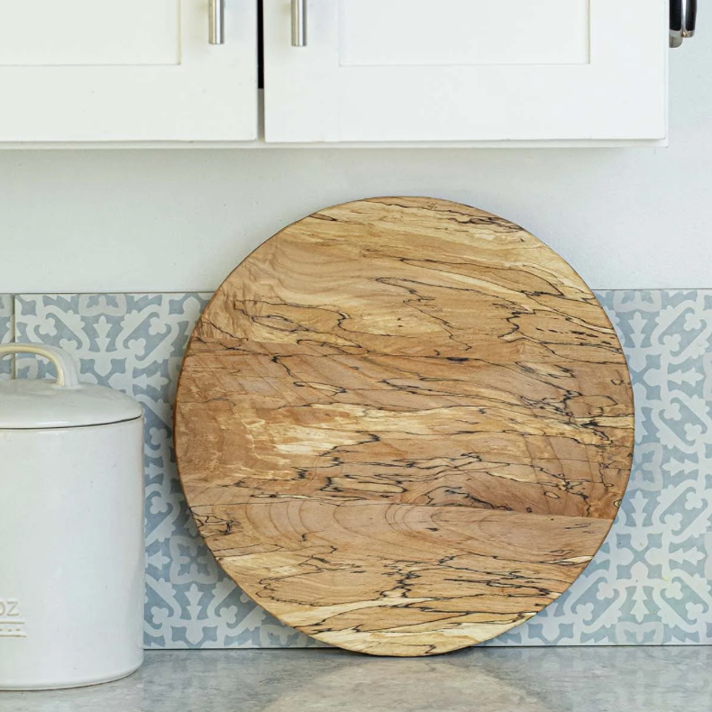 A round wooden cutting board with natural grain patterns rests against a light blue and white patterned backsplash, beside a white ceramic kitchen canister and below a white cabinet.
