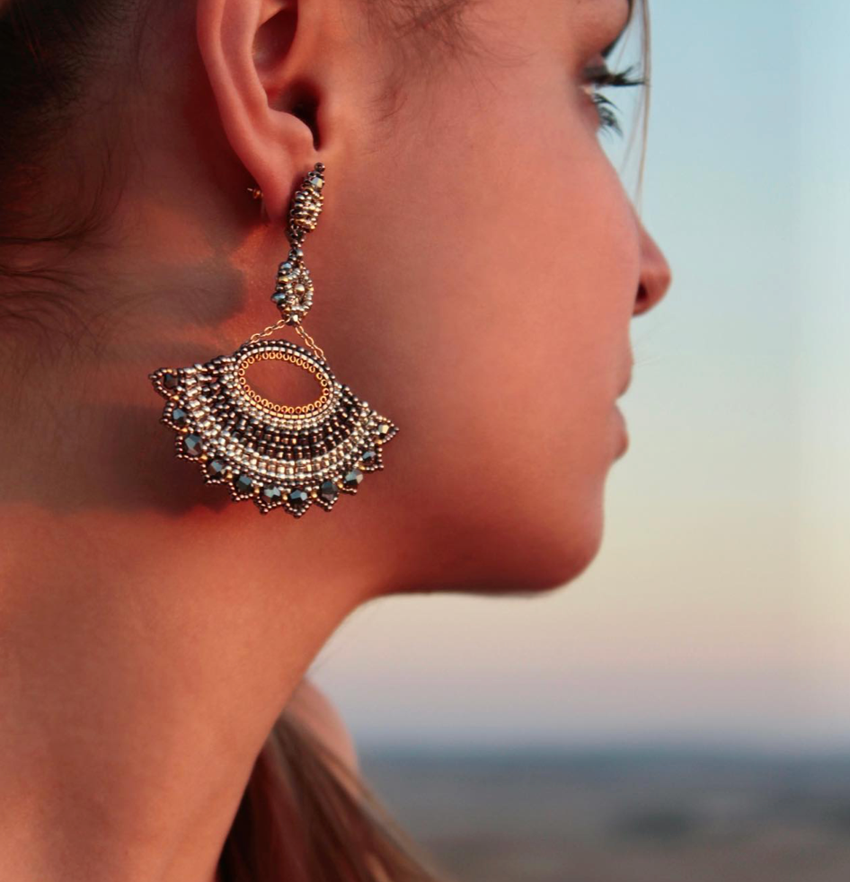 A close-up side view of a woman’s face showcasing an ornate gold and beaded chandelier earring, with a blurred outdoor background and soft, natural lighting.