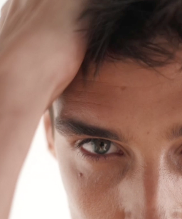 Close-up of a person with short dark hair, touching their head with one hand. Only the upper part of their face, including their intense eyes and forehead, is visible. The background is out of focus and light.