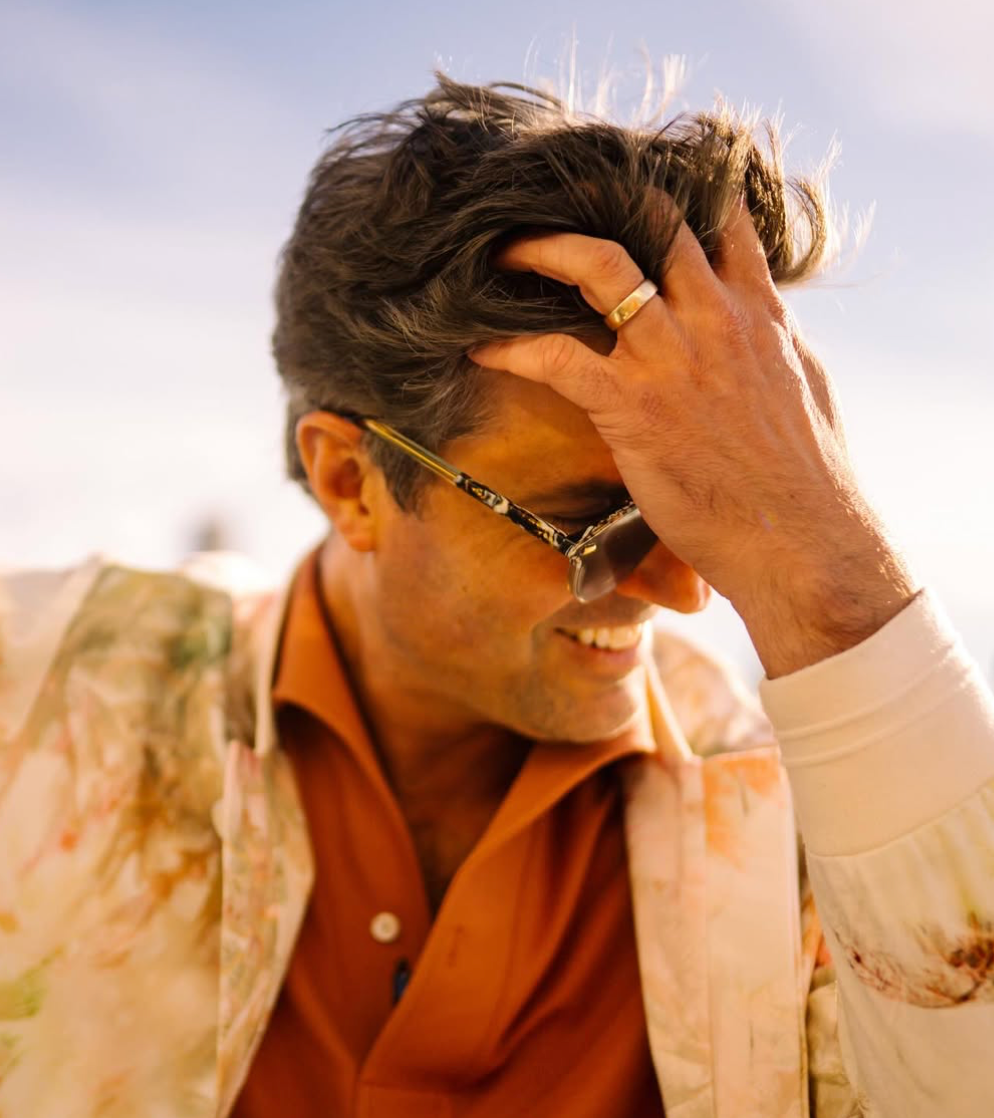 A man wearing sunglasses and an orange shirt smiles while running his hand through his hair. He is outdoors in warm sunlight, and his jacket has a colorful, light pattern.