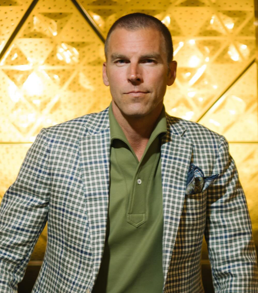 A man with short hair in a green collared shirt and checkered blazer stands in front of a gold, geometric-patterned wall, looking directly at the camera with a neutral expression.