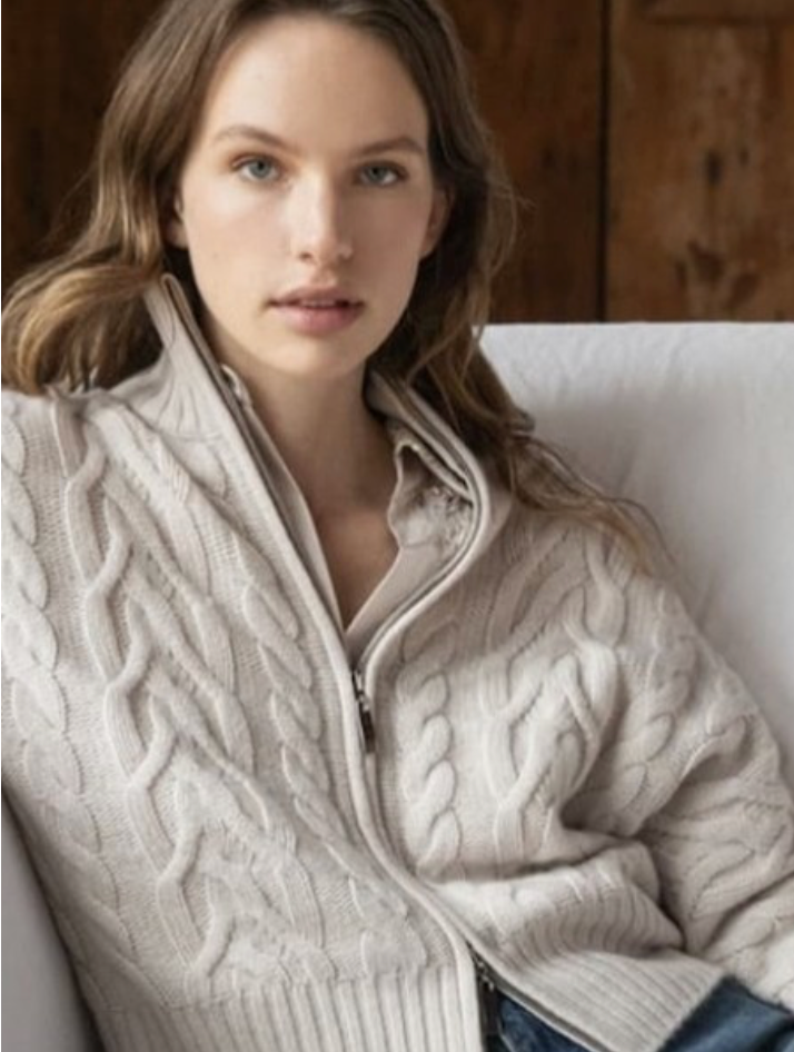 A woman with long, light brown hair sits on a white couch, wearing a light beige, cable-knit zip-up sweater over a collared shirt. She looks directly at the camera with a neutral expression.
