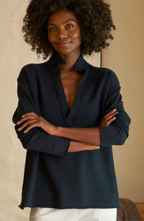 A woman with curly hair stands with arms crossed, wearing a dark long-sleeve collared top and white pants, smiling softly in a neutral indoor setting.