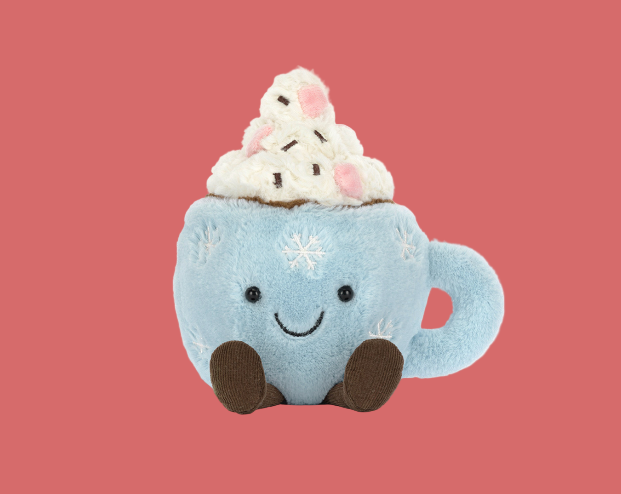 A plush toy shaped like a blue mug with a smiling face, snowflake details, and brown feet, topped with a plush swirl of whipped cream and chocolate chips, set against a solid pink background.