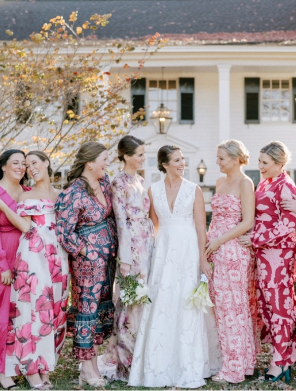 A bride in a white dress stands smiling with six bridesmaids wearing colorful, floral-patterned dresses in shades of pink, standing outdoors in front of a house.