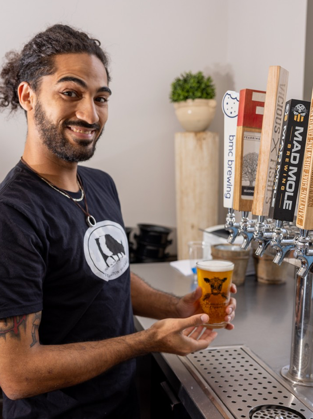 A smiling man with curly hair pours a beer from a tap at a bar, holding a pint glass with a logo. Four beer tap handles are visible, and there are bar accessories in the background.