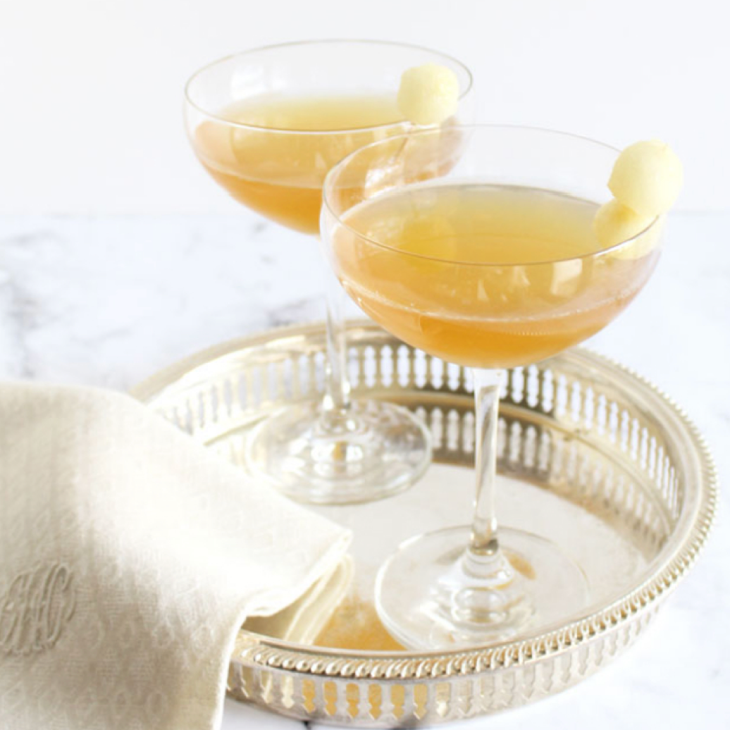 Two coupe glasses filled with a golden-brown cocktail, each garnished with a small round lemon peel, are set on a silver tray beside a folded cream-colored napkin.