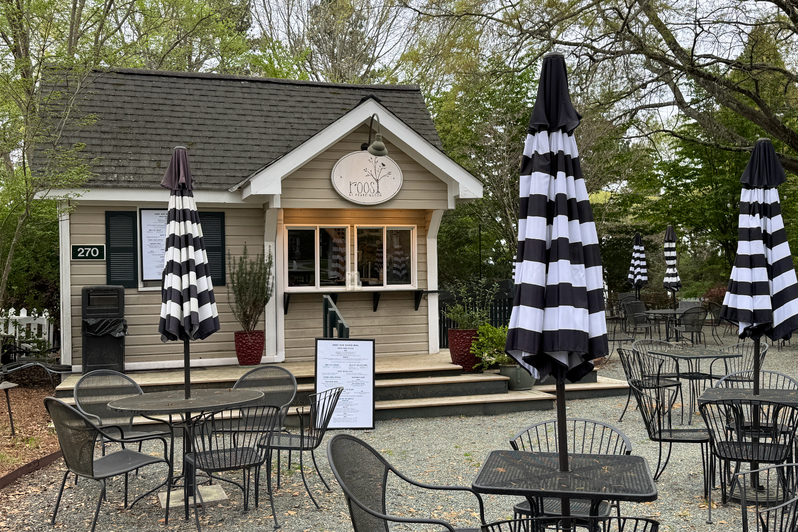 Outdoor café with black-and-white striped umbrellas, empty metal tables and chairs, and a small building with a sign reading “moo” above a takeout window. Green trees and a gravel ground surround the area.
