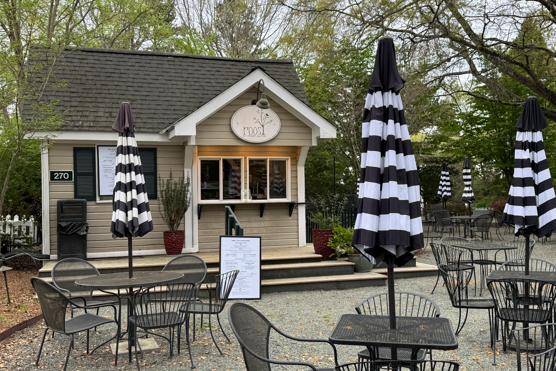 Outdoor café with black-and-white striped umbrellas, empty metal tables and chairs, and a small building with a sign reading “moo” above a takeout window. Green trees and a gravel ground surround the area.