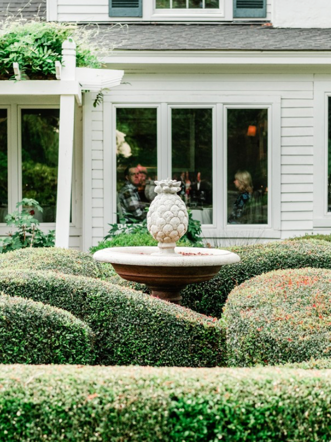 A white pineapple-shaped fountain stands in the center of neatly trimmed hedges, with a white house and large windows in the background. Two people are visible sitting inside the house.