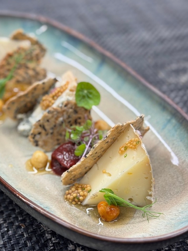 A close-up of a cheese platter with assorted cheeses, seed crackers, pickled mustard seeds, dried fruits, microgreens, and a drizzle of honey, all artfully arranged on a ceramic plate.