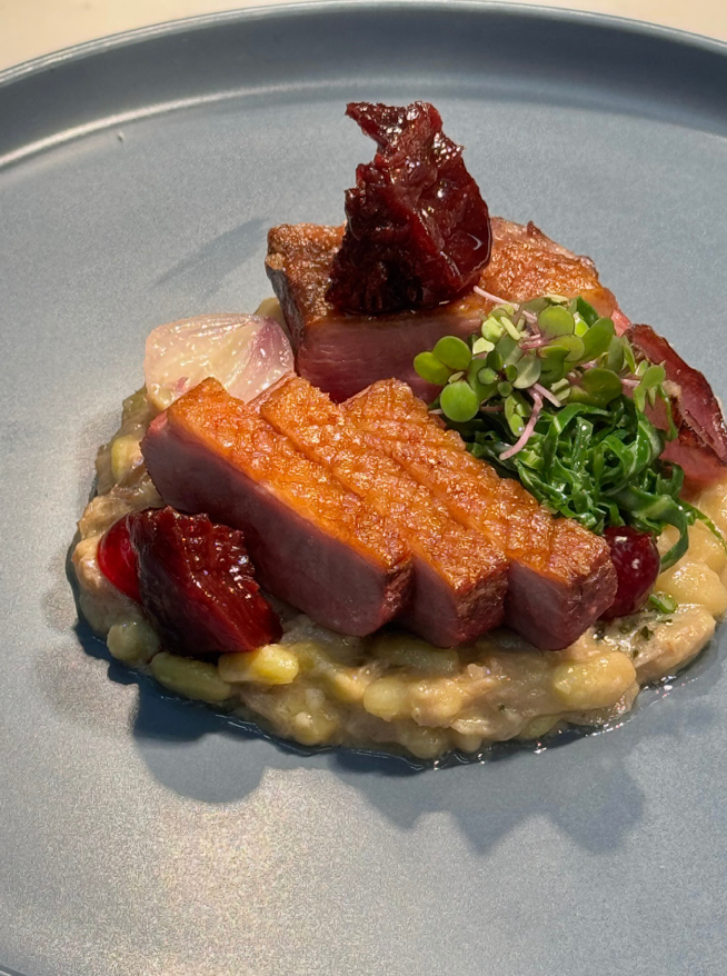 A gourmet dish featuring seared slices of duck breast atop a creamy bean puree, garnished with microgreens and glossy pieces of roasted beetroot, served on a round gray plate.