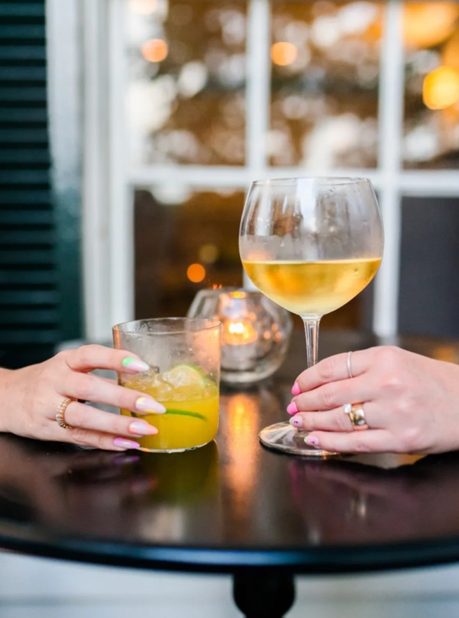 Two hands holding drinks across a round black table—a glass of orange beverage with ice and a wine glass with white wine. Candlelight and a window with greenery are blurred in the background.