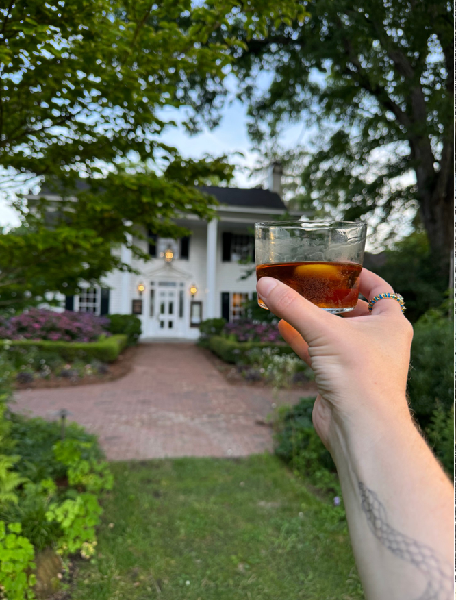 A hand holding a glass of dark beverage is raised in the foreground, with a large white house featuring columns and a garden path visible in the background. Lush greenery and trees surround the scene.