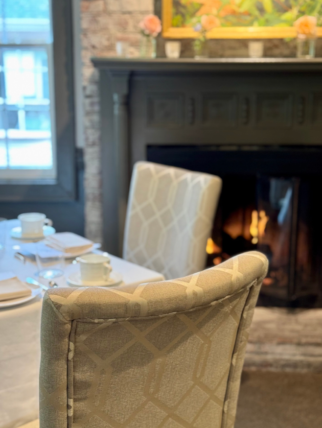A cozy dining area with cushioned chairs around a table set with white china and napkins, positioned near a lit fireplace and a dark mantel decorated with small vases of flowers.