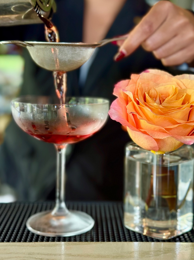 A person strains a red cocktail into a coupe glass beside an orange rose in a small glass vase on a bar counter.