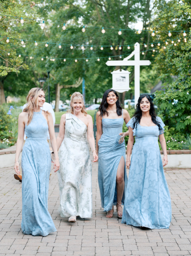 Four women in elegant, light blue and white dresses walk together outdoors on a stone path, smiling and holding hands, with string lights and greenery in the background.