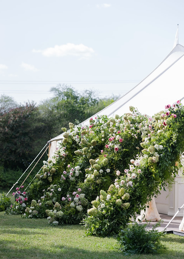 A white tent with a grand entrance draped in lush greenery and blooming white and pink flowers, set outdoors on a grassy lawn under a bright sky.