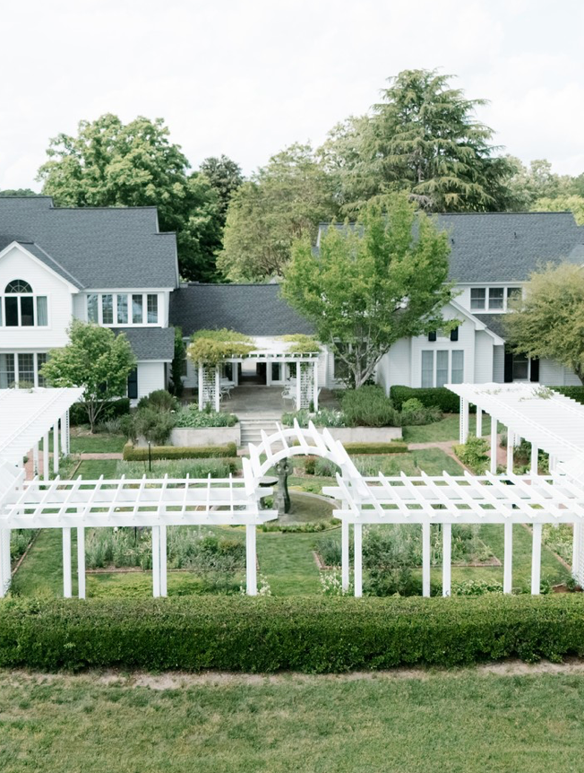 A large, elegant white house with dark gray roofs stands behind a manicured garden featuring white wooden pergolas, green hedges, and neatly arranged garden beds, surrounded by tall trees on a bright day.