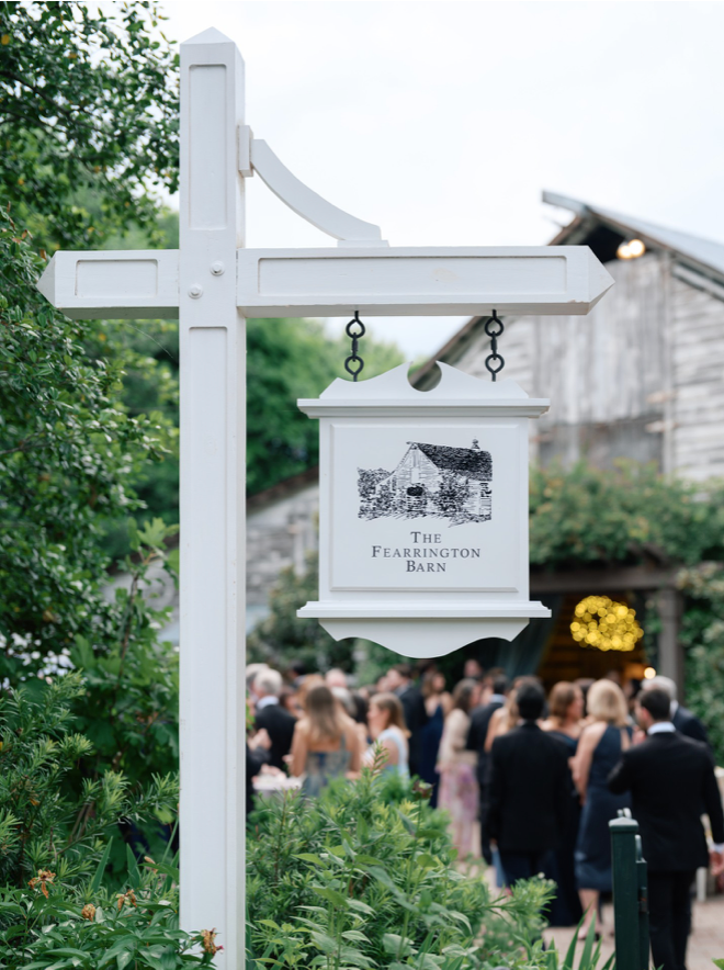 A white signpost for The Fearrington Barn stands in the foreground, with a crowd of formally dressed people gathered near a rustic barn in the background, surrounded by greenery.