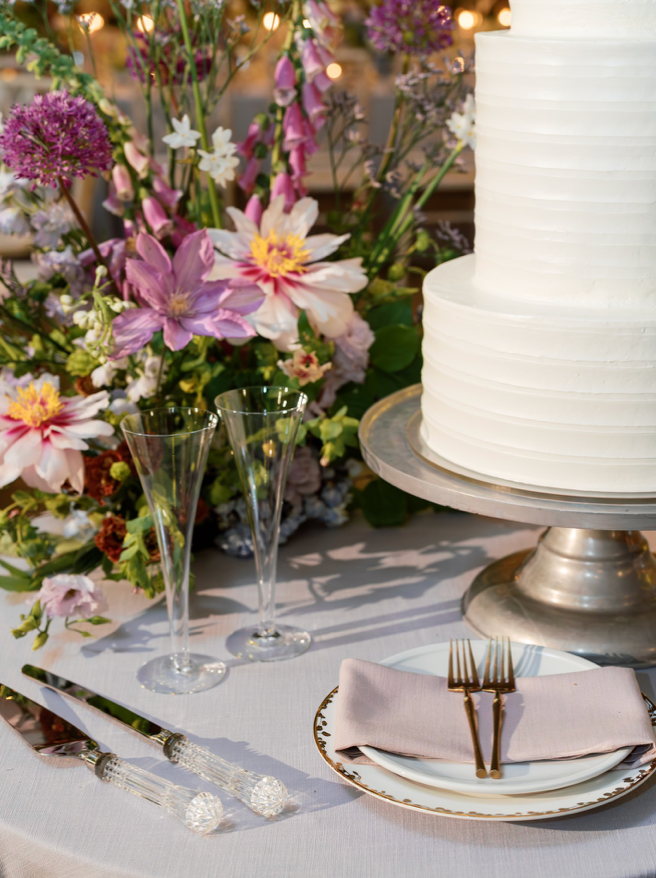 A white tiered cake on a silver stand is displayed next to two champagne flutes, a plate with pink napkin and gold utensils, and colorful flowers on a white tablecloth.