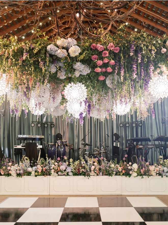 A stage decorated with musical instruments and surrounded by flowers. Above the stage hangs a lush floral arrangement with pink, purple, and white flowers, greenery, and glowing lights, under a wooden ceiling with string lights. The floor is checkered.