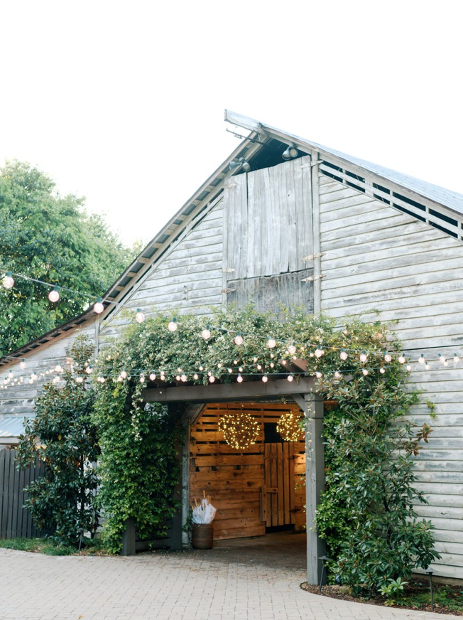 A rustic wooden barn decorated with string lights and greenery, with open doors revealing more lights inside. The barn is surrounded by trees and bushes, and the scene feels festive and inviting.