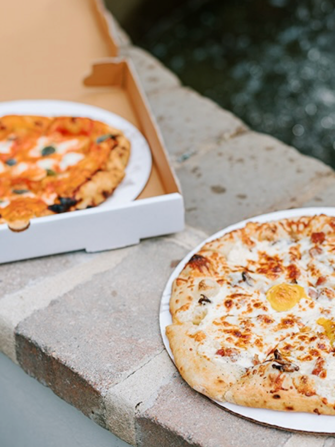 Two pizzas on white plates rest on a stone ledge outdoors; one pizza is in an open cardboard box, while the other is topped with cheese and egg slices, placed closer to the camera.