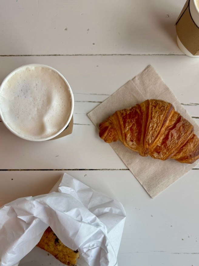 A cup of frothy coffee, a croissant on a napkin, and a partially unwrapped cookie on a white wooden table, with part of another coffee cup visible in the corner.
