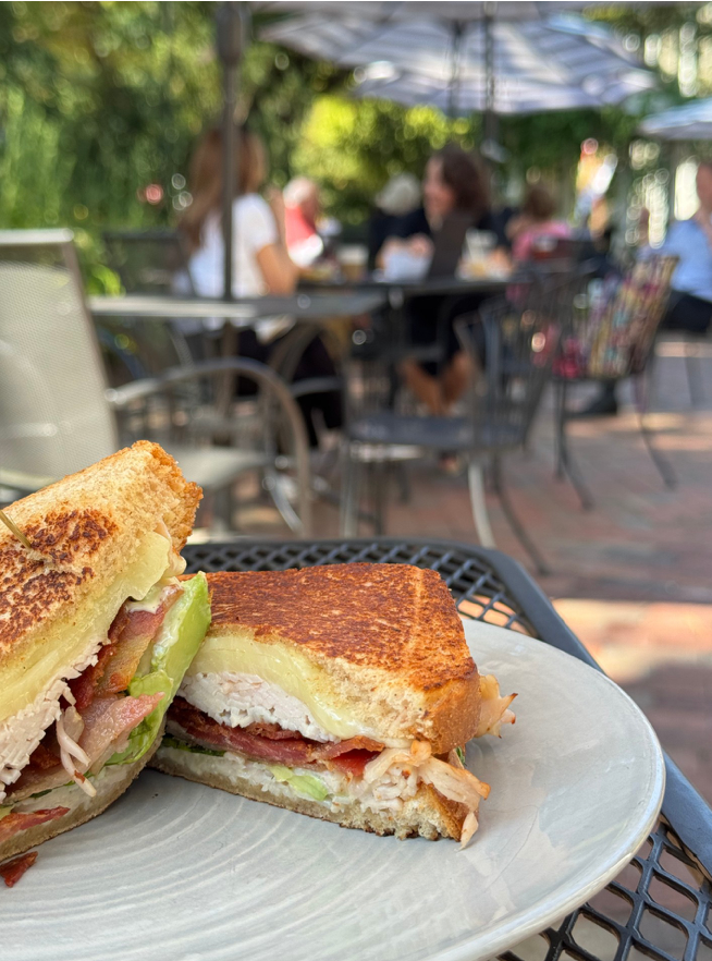 A close-up of a grilled sandwich with turkey, bacon, cheese, and avocado on a plate, set on an outdoor patio table with blurred people sitting in the background.