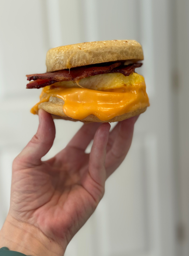 A hand holds up a breakfast sandwich with bacon, a fried egg, and melted cheddar cheese on an English muffin, against a blurred white background.