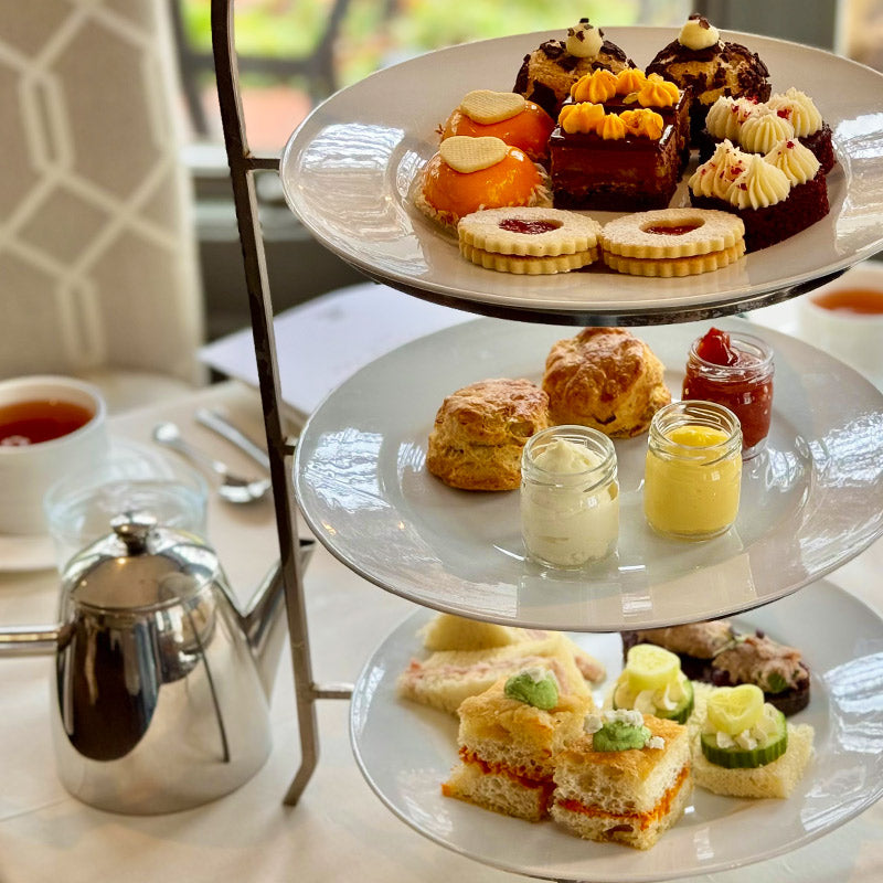 A three-tiered tray with assorted afternoon tea treats: cakes and pastries on the top, scones with jam and cream in the middle, and finger sandwiches on the bottom. A teapot and cup are placed beside the stand.