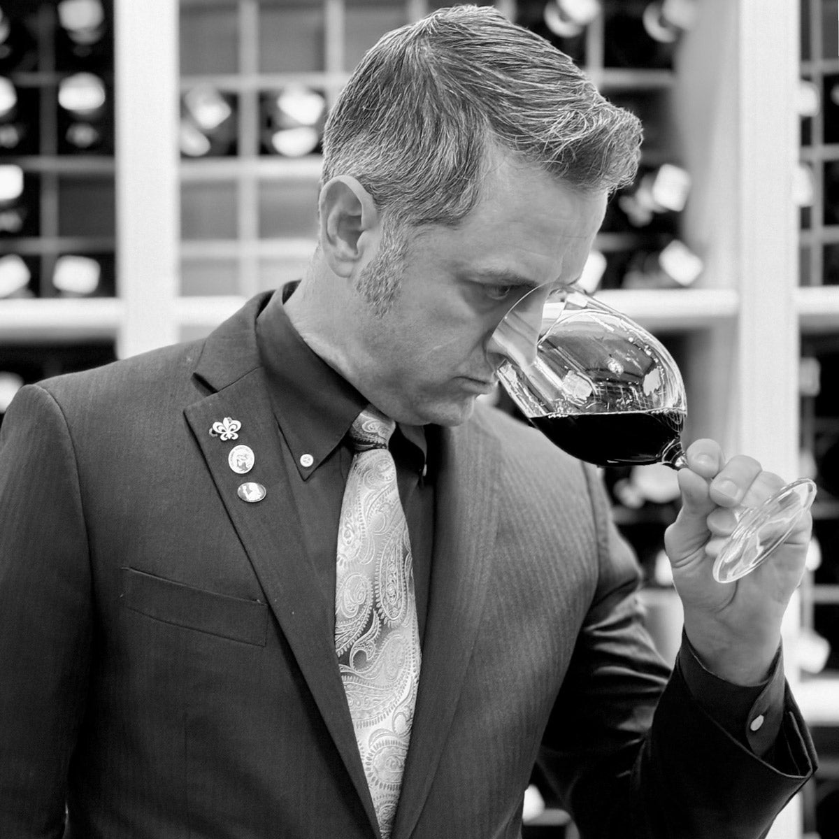 A man in a suit smells a glass of red wine, thoughtfully evaluating its aroma. Shelves of wine bottles are visible in the background. The photo is in black and white.