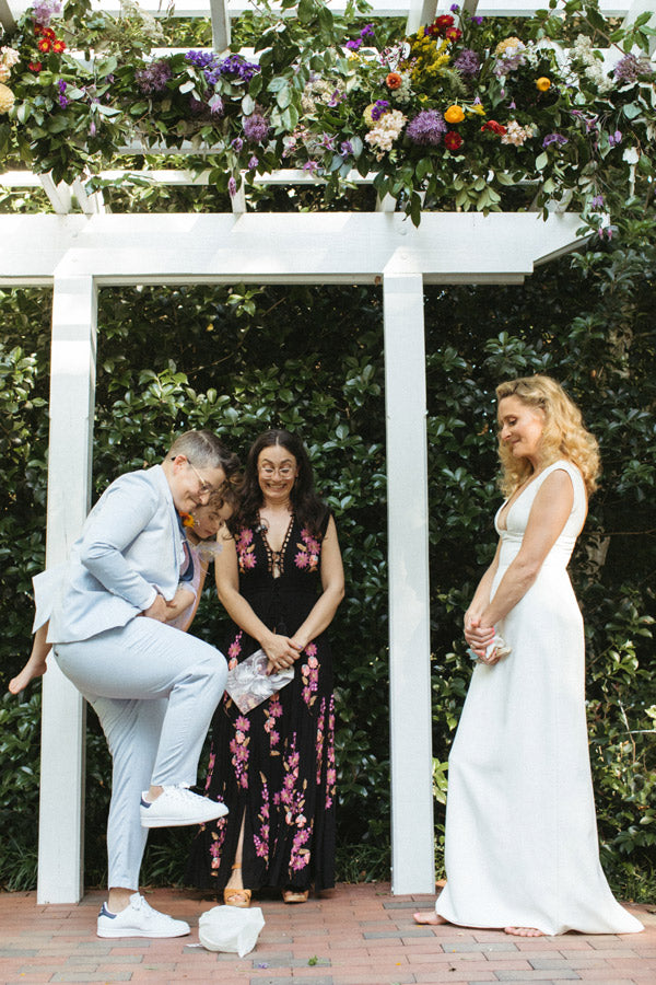 A person in a light suit steps on a glass with a child in their arms, while another person in a white dress smiles. Two people stand behind them under a floral arch, outdoors.