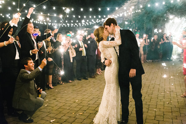 A bride and groom kiss under string lights at night, surrounded by guests holding sparklers and dressed in formal attire, celebrating outdoors on a stone-paved area.