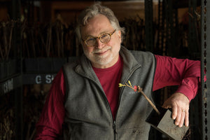 An older man with gray hair and glasses, in a red shirt and gray vest, smiles as he holds a small plant before shelves of greenery, capturing the inviting charm of a FEARRINGTON VILLAGE St. Innocent Wine Dinner.
