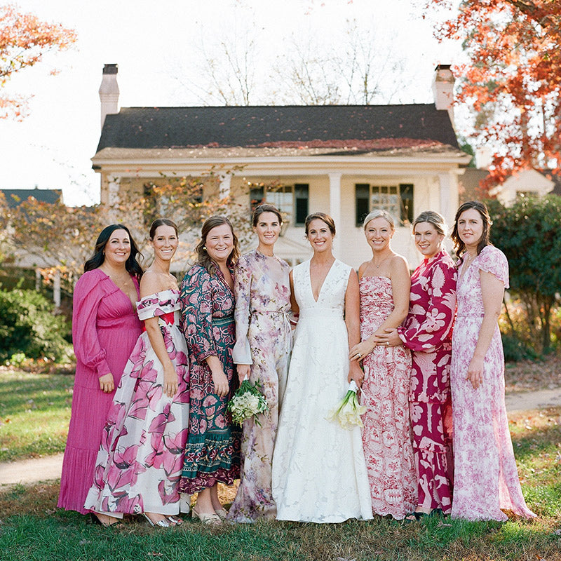 Eight women, including a bride in a white dress, stand together outdoors in front of a house with autumn trees. The bridesmaids wear pink or floral dresses and are smiling at the camera.