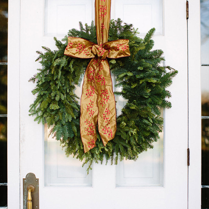 A green pine wreath with a gold ribbon decorated with red patterns hangs on a white door with a brass handle.