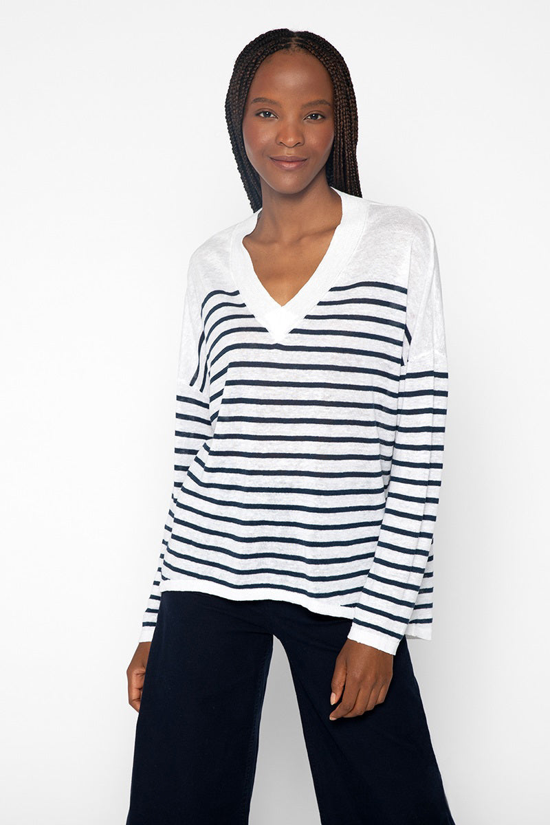 A person with long braided hair is smiling while wearing a white KINROSS CASHMERE - EASY VEE LINEN SWEATER with horizontal black stripes from the brand KINROSS. They are also wearing dark pants and standing against a plain white background.