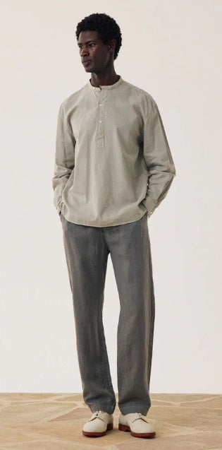 A man stands against a plain background wearing a light gray long-sleeve henley shirt, HARTFORD - LINEN JOGGY PANTS IN ARMY, and light-colored shoes, with his hands in his pockets.