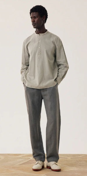 A man stands against a plain background wearing a light gray long-sleeve henley shirt, HARTFORD - LINEN JOGGY PANTS IN ARMY, and light-colored shoes, with his hands in his pockets.