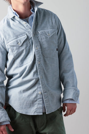 Dressed in the RELWEN Utility Workshirt in cornmeal herringbone layered over a blue and white striped shirt and paired with green pants, the individual stands with relaxed hands by their sides, capturing the effortless sophistication of luxury workwear. The person’s head is partially out of frame.
