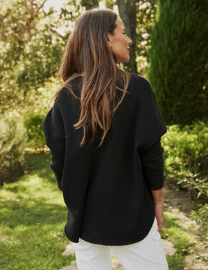 A person with long, wavy blonde hair is standing outdoors near a large tree, wearing the FRANK & EILEEN ANNA LONG-SLEEVE CAPELET in navy over a white shirt and blue pants. The background showcases green foliage and a stone path. The person is smiling and gazing to the side.
