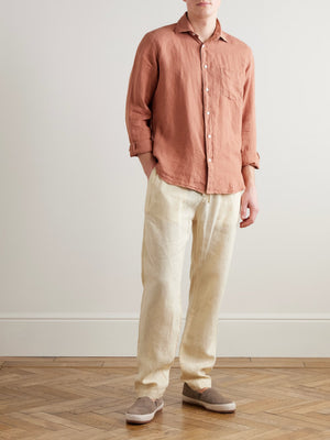 A man, seen from the back with hands in pockets, wears the HARTFORD - PAUL LINEN SHIRT IN FADED COPPER with rolled-up sleeves, paired with light pants.
